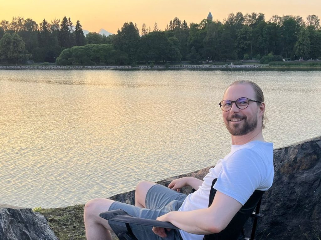Teemu Lari sitting by a lake during sunset, wearing a white shirt and glasses, smiling at the camera.